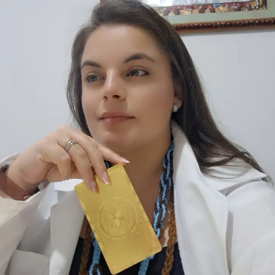 Priscila de blazer branco e guias no pescoço, segurando uma carta de tarot dourada