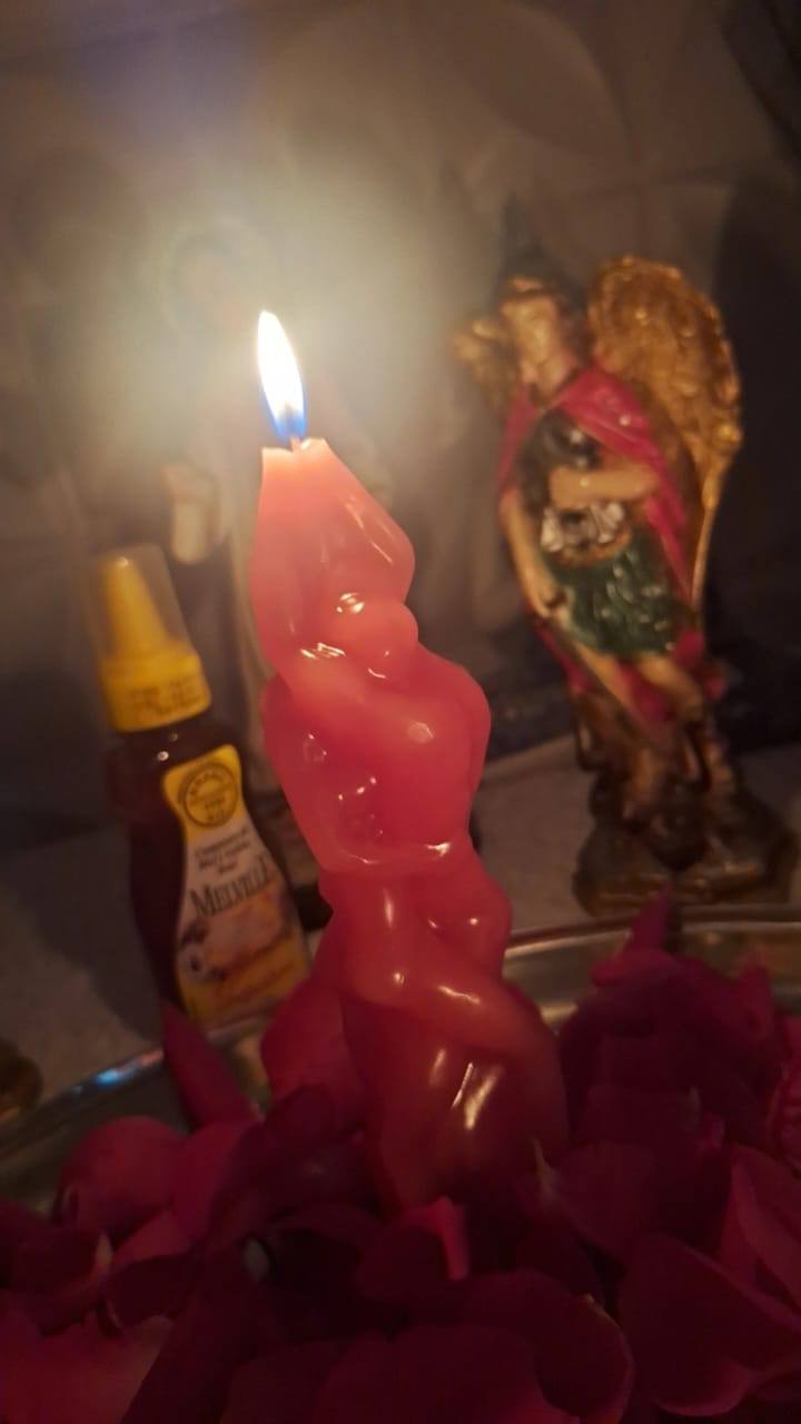 Vela vermelha em forma de casal abraçado, acesa sobre pétalas de rosa, com mel e a imagem de um anjo ao fundo